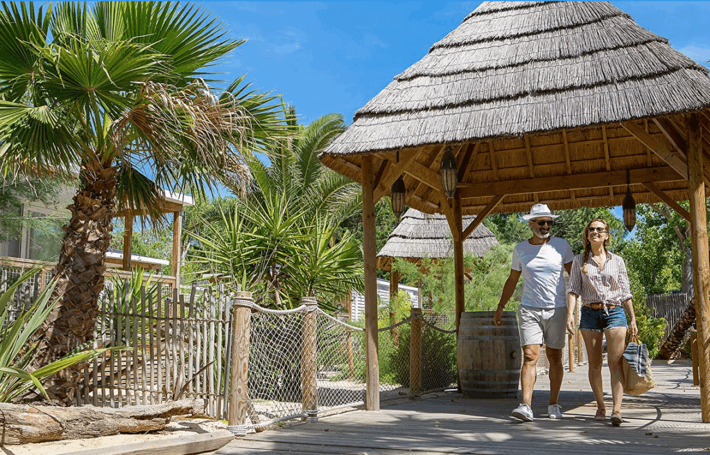 Paillote en bois et roseau de Camargue vue sur une bordure de palmiers avec à droite une paillote en toit de roseau de Camargue conçue par Ekolinea, avec un homme et une femme qui se tiennent la main en souriant.