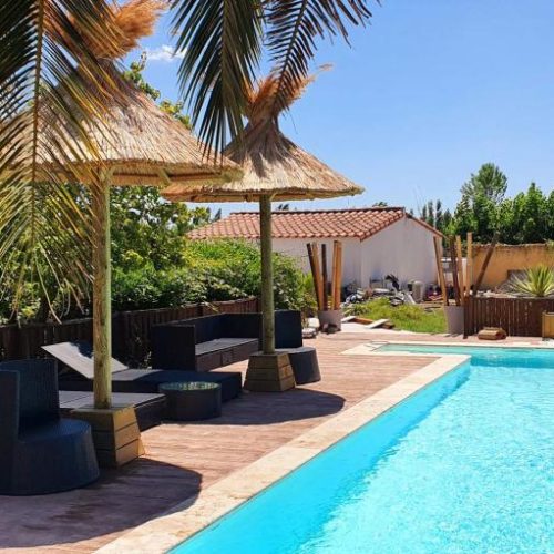 Vue derrière des branches de palmier d'une piscine et de deux parasols exotiques EKOLINEA avec des transats